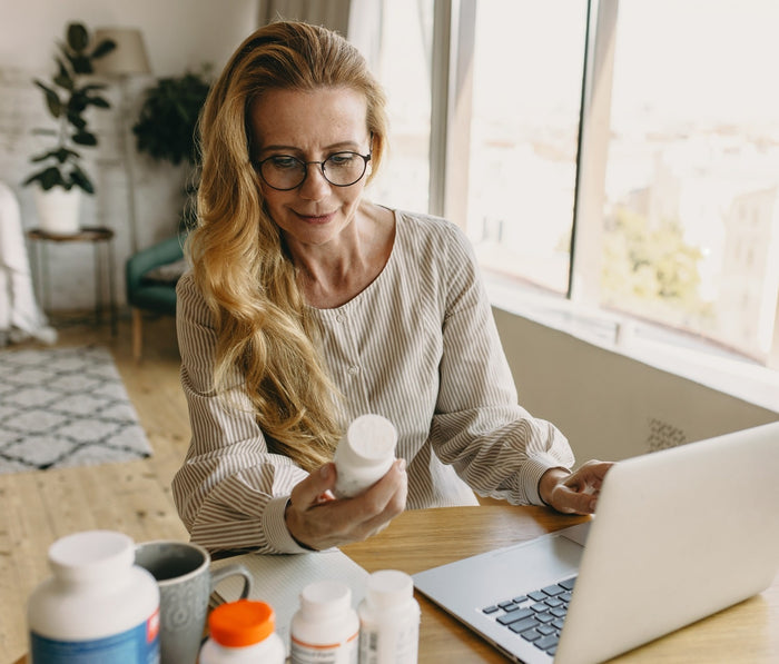 Eine ältere Dame sitzt in ihrem Wohnzimmer vor einem Laptop und hält eine Nahrungsergänzungsmitteldose in der Hand.