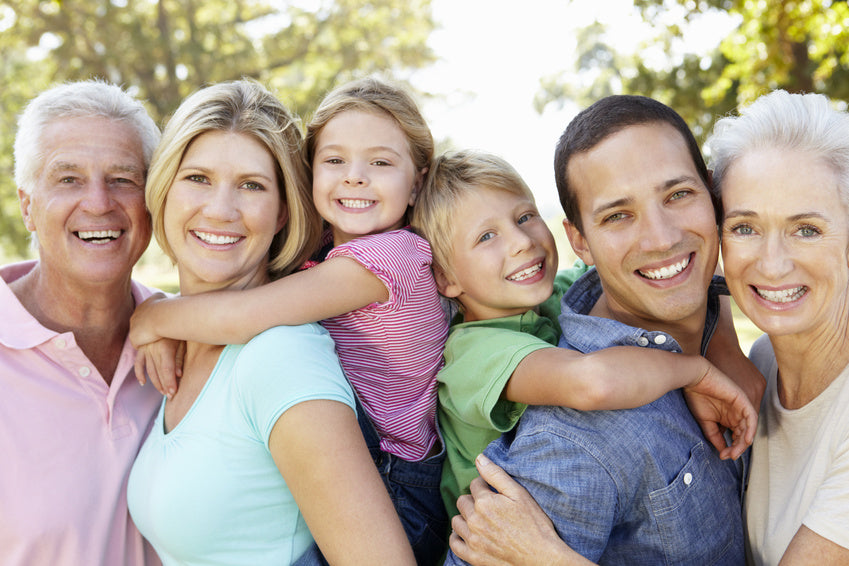 Eine Familie bestehend aus Großvater, Großmutter, Mutter, Vater, Kind (m) und Kind (w) sind in einem Garten und lächeln zusammen in die Kamera.