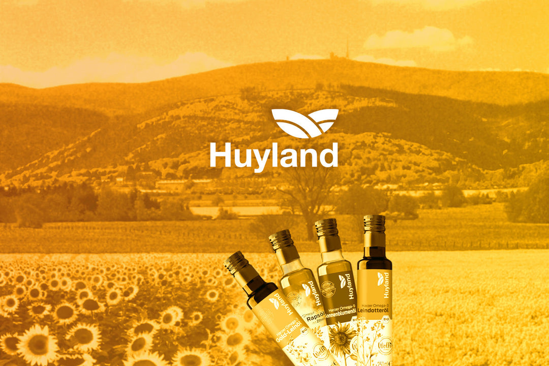 4 Huyland Öle stehen nebeneinander vor einem Hintergrund mit Raps und Sonnenblumenfeldern. Im Zentrum des Bildes sieht man das Huyland Markenbild und den Namen der Marke - Huyland.